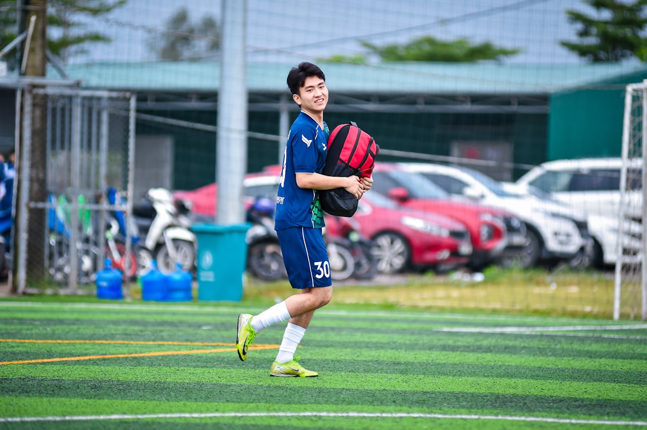 A smiling young football player carrying sports gear on an outdoor field in Hanoi, Vietnam.